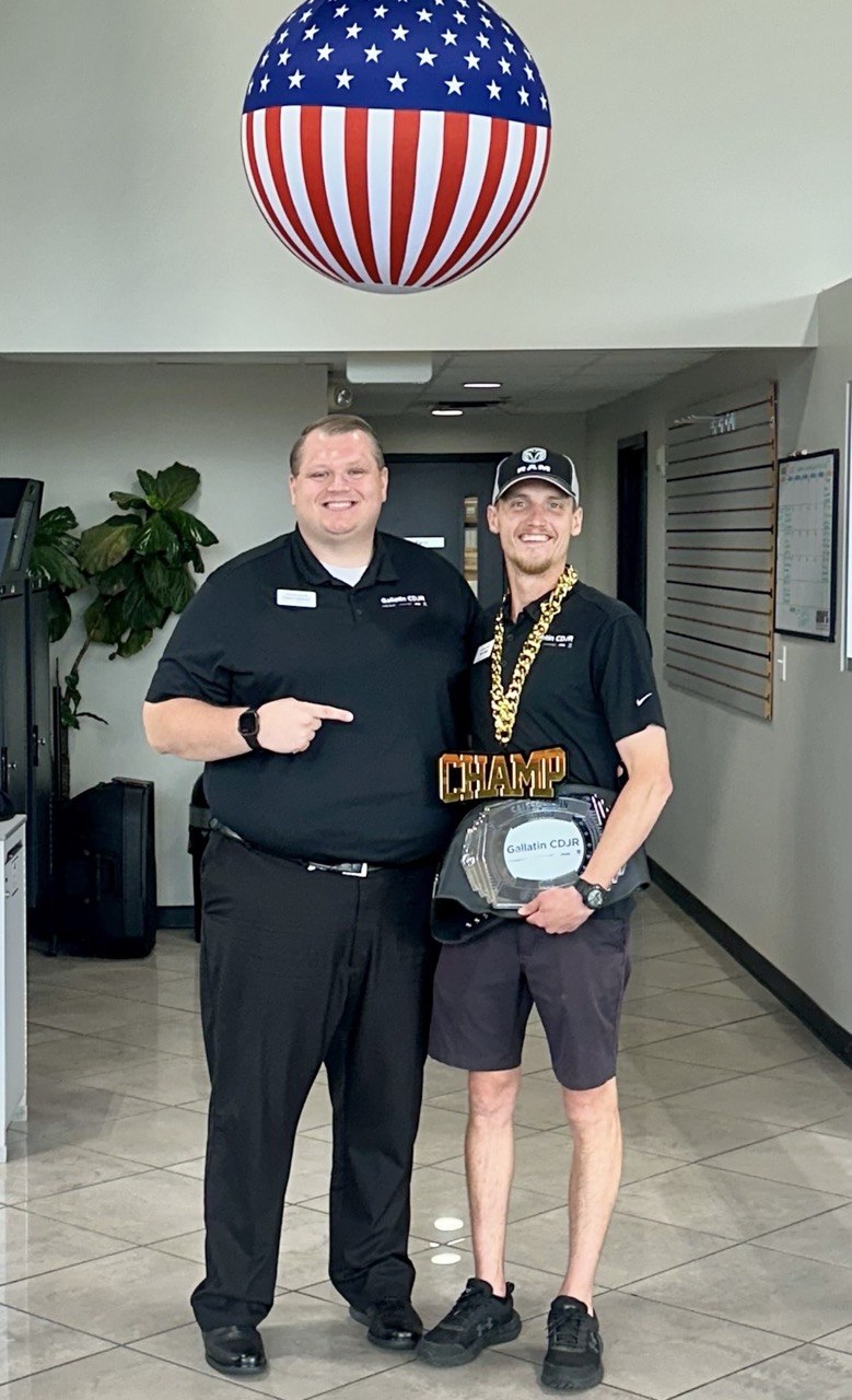 A teammate in a Gallatin CDJR polo points to a colleague in a RAM cap holding the Salesperson of the Month belt and a gold CHAMP chain