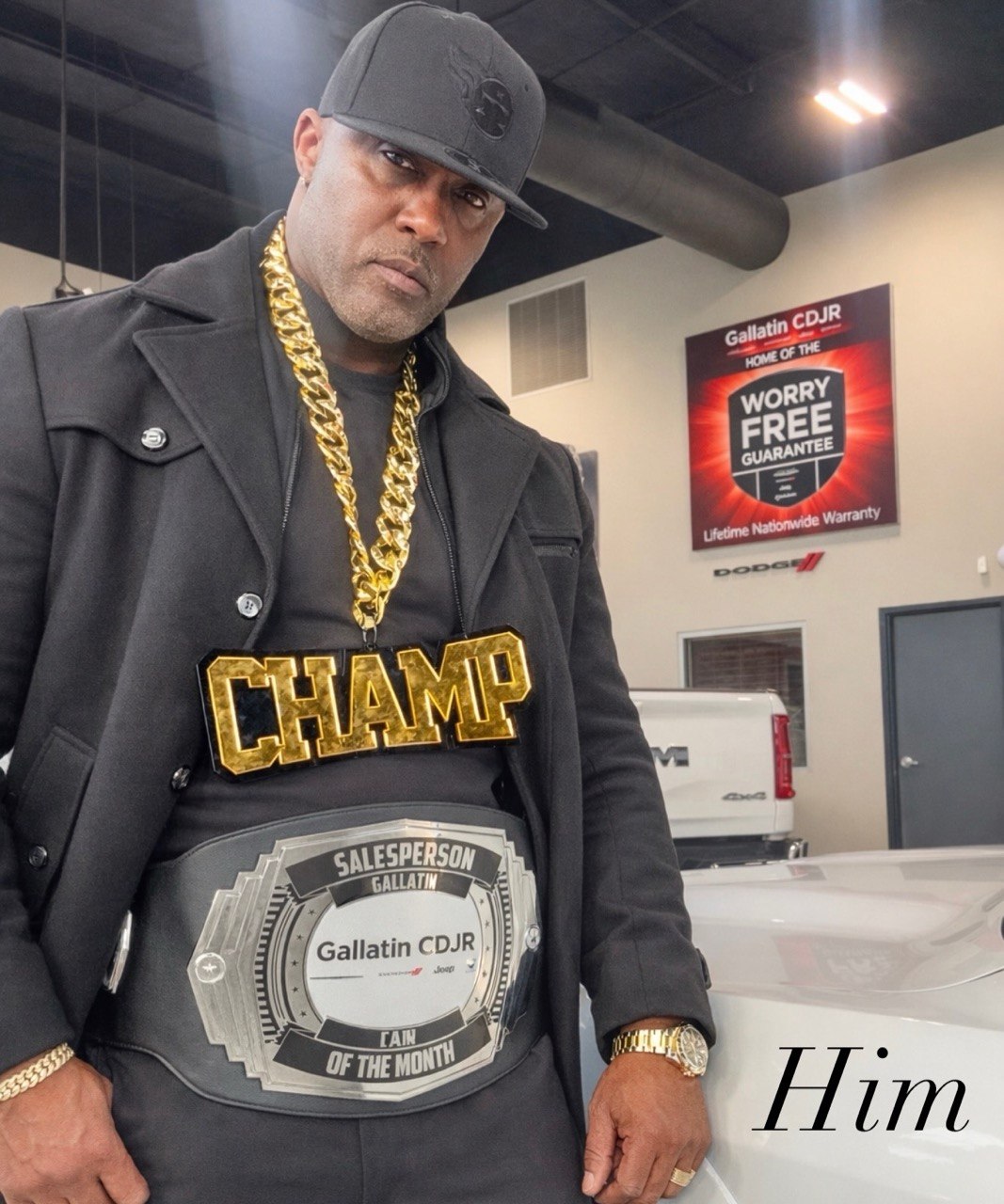 A man in an all-black outfit and black cap wears the Salesperson of the Month belt and a gold CHAMP chain in the showroom