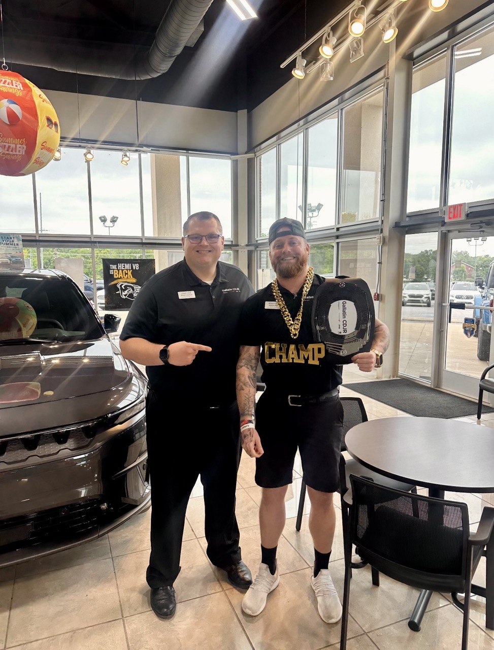 A bearded man in a CHAMP shirt holds the belt in front of a 'THE HEMI V8 IS BACK' sign and a beach-ball summer promo