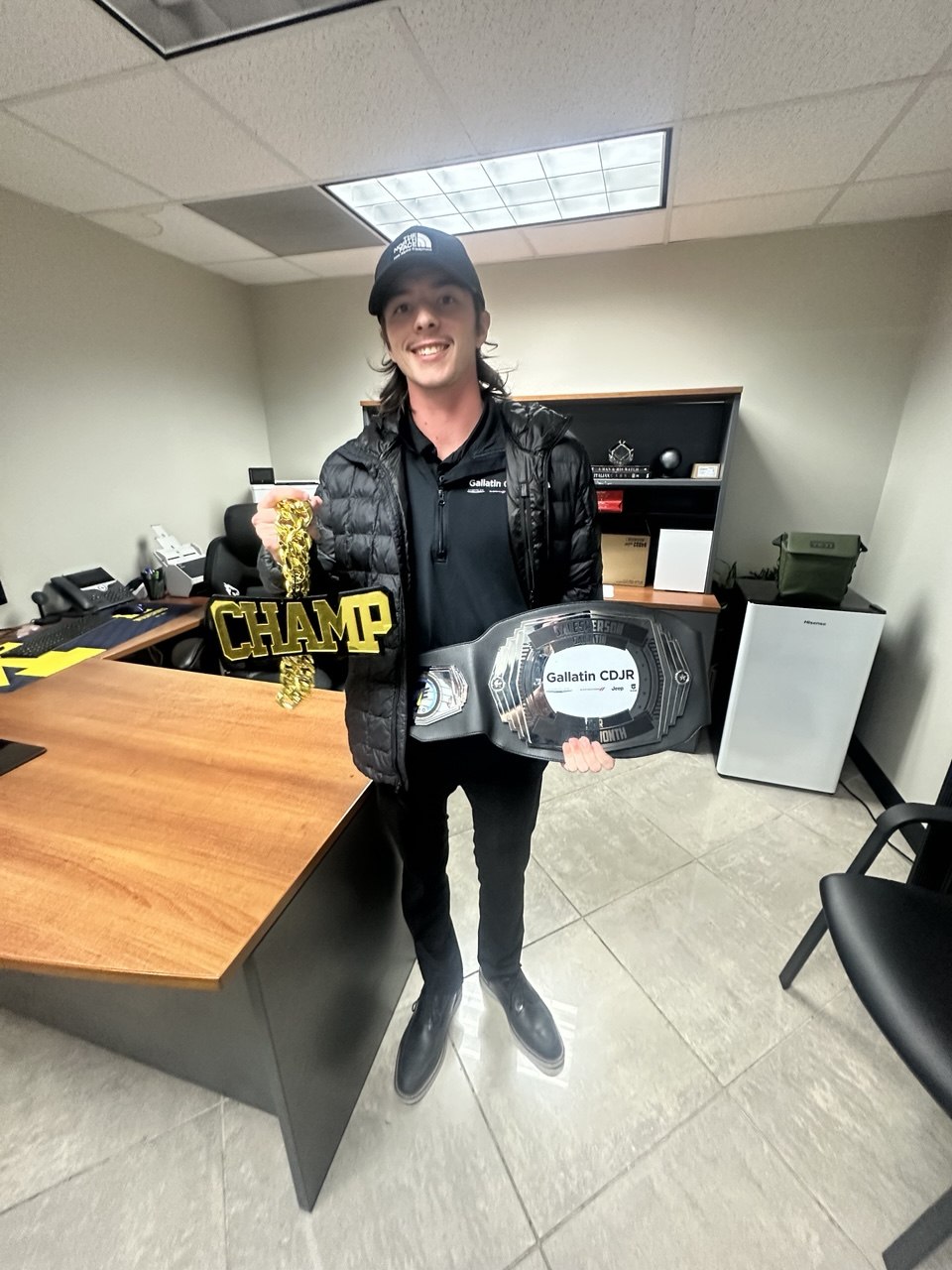 A young man with a mullet and North Face cap holds a gold CHAMP chain and the championship belt
