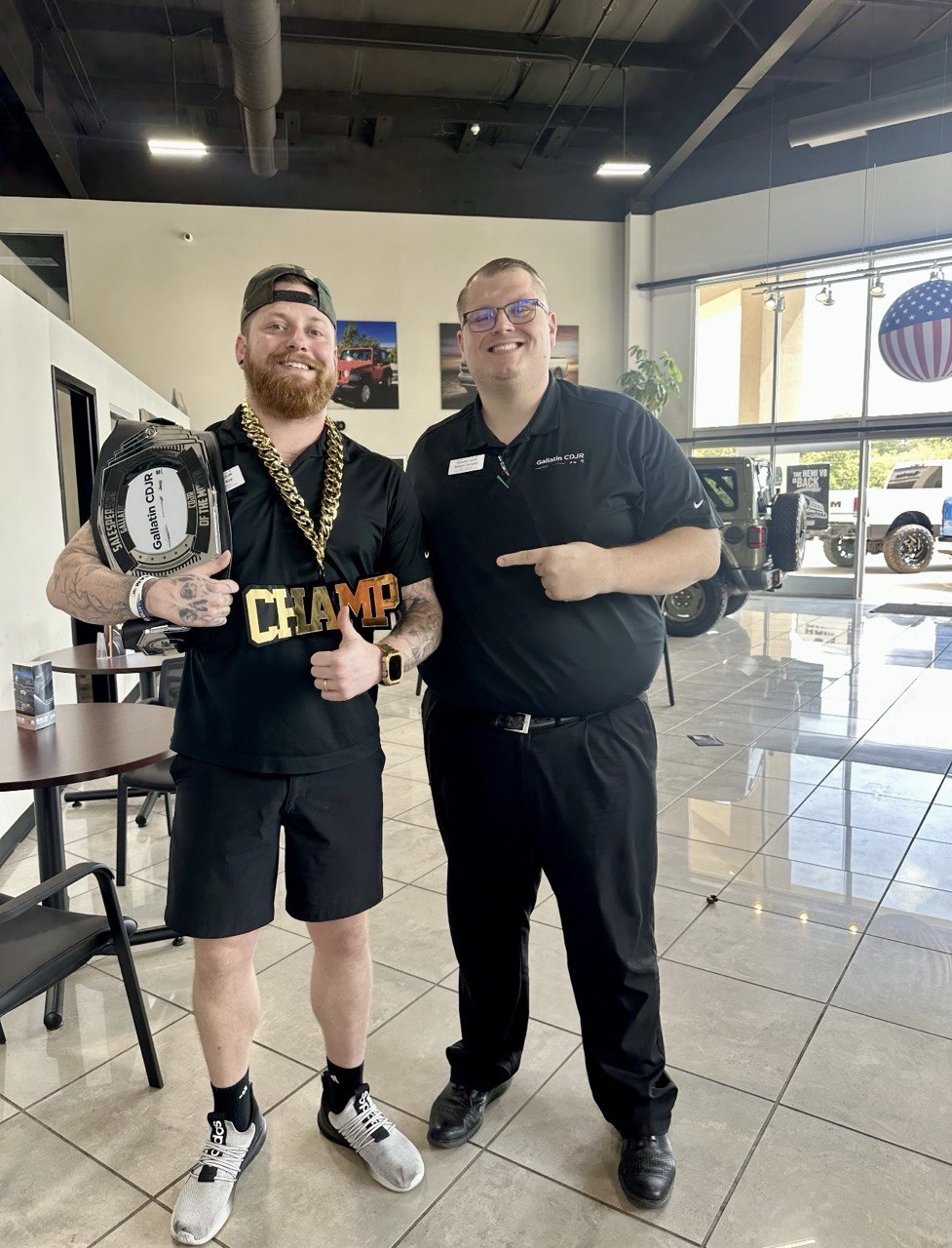 A man with a red beard and backwards cap in a black CHAMP shirt holds the belt and gives a thumbs-up while a colleague points